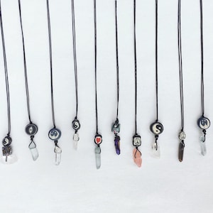 May include: A collection of 14 necklaces with various charms, including peace signs, yin yang symbols, hearts, and crystals. The necklaces are all made with black string and have a simple, minimalist design.