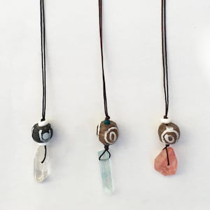 May include: Three handmade necklaces with natural stone pendants. Each necklace features a unique stone pendant, including clear, light blue, and pink crystals. The beads are brown and white with a spiral design, suspended from a black or brown cord.