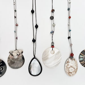 May include: Six necklaces with pendants of various shapes and materials hang against a white background. The pendants include a teardrop shape, a circular shape, and a shell shape. The necklaces are strung on black cords with colorful beads.