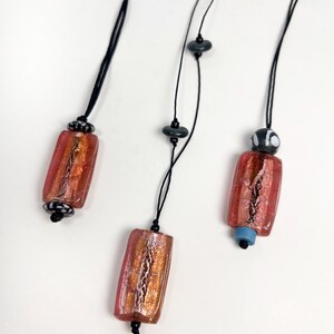 May include: Three necklaces with red and gold glass pendant beads and black cord. The beads have a metallic sheen and are rectangular in shape.