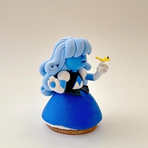 Op de afbeelding: Een handgemaakt beeldje van een blauw personage met lichtblauw haar, een donkerblauwe jurk en witte handschoenen. Het personage houdt een kleine gele vogel vast. Het beeldje staat op een houten voet.