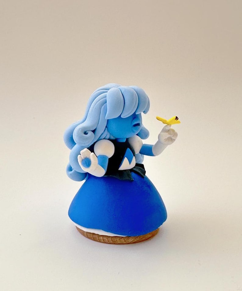 Op de afbeelding: Een handgemaakt beeldje van een blauw personage met lichtblauw haar, een donkerblauwe jurk en witte handschoenen. Het personage houdt een kleine gele vogel vast. Het beeldje staat op een houten voet.