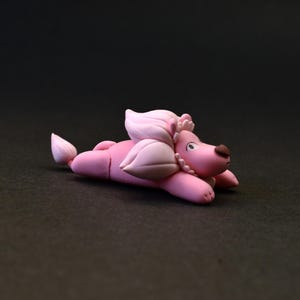 Op de afbeelding: Een roze poedelfiguur, liggend, met een lichtroze manen en staart. De poedel heeft een bruine neus en is versierd met een parelketting. Het beeldje staat op een donkere achtergrond.