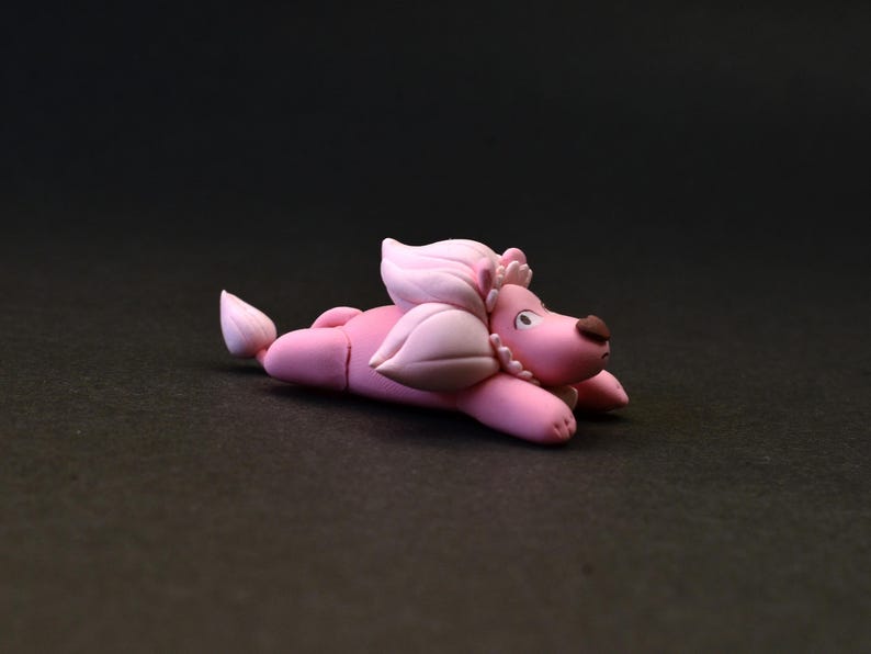 Op de afbeelding: Een roze poedelfiguur, liggend, met een lichtroze manen en staart. De poedel heeft een bruine neus en is versierd met een parelketting. Het beeldje staat op een donkere achtergrond.
