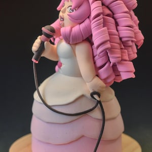 Op de afbeelding: Handgemaakte figuur van een personage met roze haar, met een microfoon. De figuur draagt een witte en roze jurk, staand op een houten voet. De microfoon is zwart met een rode accent.