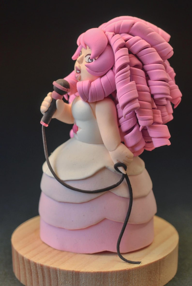 Op de afbeelding: Handgemaakte figuur van een personage met roze haar, met een microfoon. De figuur draagt een witte en roze jurk, staand op een houten voet. De microfoon is zwart met een rode accent.