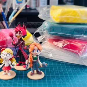 May include: Three handcrafted clay figurines featuring a tall character in a red suit with a staff, a small blonde character in a white dress, and a character with braided orange hair holding tools, alongside several blocks of colored modeling clay.