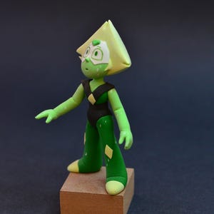 Op de afbeelding: Handgemaakte figuur van een groen personage met een driehoekig hoofd, gekleed in een zwart bodysuit en groene broek met gele diamantaccenten. De figuur staat op een klein houten blok tegen een donkere achtergrond.