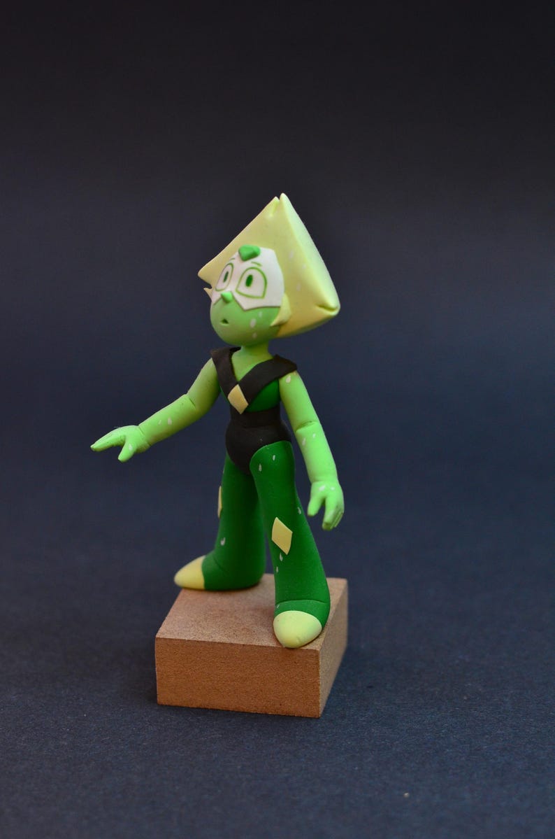 Op de afbeelding: Handgemaakte figuur van een groen personage met een driehoekig hoofd, gekleed in een zwart bodysuit en groene broek met gele diamantaccenten. De figuur staat op een klein houten blok tegen een donkere achtergrond.