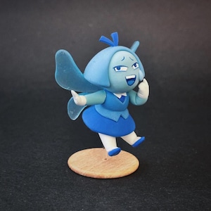 Op de afbeelding: Handgemaakt beeldje van een stripfiguur in blauwtinten. Het figuur heeft een lichtblauwe kop met een donkerblauw haaraccessoire, een donkerblauwe jurk en witte handschoenen. Het staat op een kleine, ronde houten basis met een vrolijke uitdrukking.
