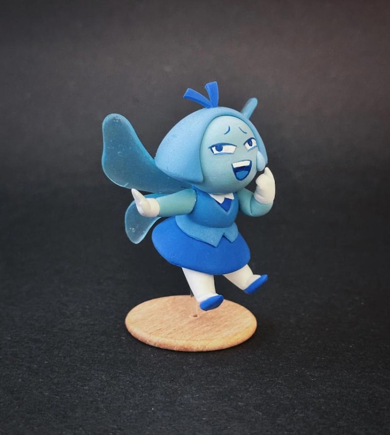 Op de afbeelding: Handgemaakt beeldje van een stripfiguur in blauwtinten. Het figuur heeft een lichtblauwe kop met een donkerblauw haaraccessoire, een donkerblauwe jurk en witte handschoenen. Het staat op een kleine, ronde houten basis met een vrolijke uitdrukking.