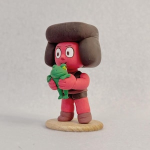 Op de afbeelding: Handgemaakte kleifiguur van een personage met een rode huid, bruin haar en een bruin outfit, die een groene kikker vasthoudt. De figuur staat op een kleine, ronde houten basis. Het personage heeft grote witte ogen.