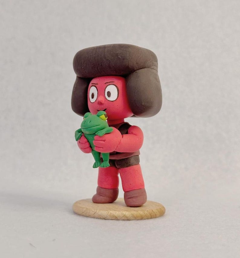 Op de afbeelding: Handgemaakte kleifiguur van een personage met een rode huid, bruin haar en een bruin outfit, die een groene kikker vasthoudt. De figuur staat op een kleine, ronde houten basis. Het personage heeft grote witte ogen.