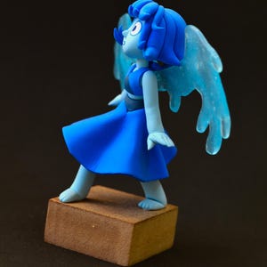 Op de afbeelding: Blauw beeldje van een personage met blauw haar, een blauwe jurk en doorschijnende blauwe vleugels. Het beeldje staat op een kleine, bruine, rechthoekige basis. Het personage kijkt omhoog en naar links.