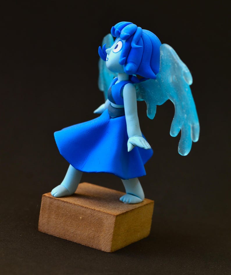 Op de afbeelding: Blauw beeldje van een personage met blauw haar, een blauwe jurk en doorschijnende blauwe vleugels. Het beeldje staat op een kleine, bruine, rechthoekige basis. Het personage kijkt omhoog en naar links.