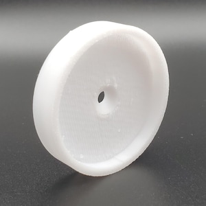 May include: A white, circular object with a hole in the center. The object has a rounded edge and a textured surface. The item appears to be made of a solid, opaque material. The object is likely a component or part of a larger product.