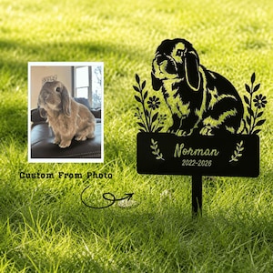 May include: A black metal garden stake featuring a silhouette of a lop-eared rabbit surrounded by floral stems. Below the rabbit, a rectangular plaque displays the name Norman and the years 2022-2026, accompanied by a small photo of a brown rabbit.