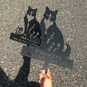 May include: Black metal cat memorial garden stake with the name "SNUGGLES" and dates March 2002 - January 2021. The cat silhouette is sitting with floral accents.