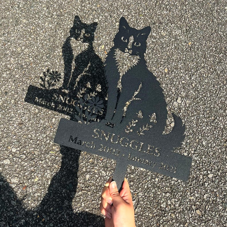 May include: Black metal cat memorial garden stake with the name "SNUGGLES" and dates March 2002 - January 2021. The cat silhouette is sitting with floral accents.