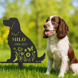 以下が含まれることがあります： 犬のシルエット、名前「MILO」、年号「2008-2025」、蝶と花の模様が施された黒い金属製のガーデンステーク。ステークは、草地に座っている茶色と白の犬の隣に配置されています。