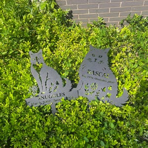 May include: Two black metal cat silhouette garden stakes. One stake reads "Snuggles" with dates, the other reads "Rascal" with dates. Both stakes are placed in a bed of green foliage.