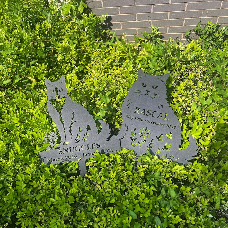 May include: Two black metal cat silhouette garden stakes. One stake reads "Snuggles" with dates, the other reads "Rascal" with dates. Both stakes are placed in a bed of green foliage.