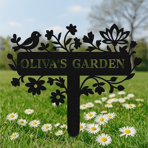 May include: Black metal garden stake with the text "OLIVA'S GARDEN". The stake features a bird, flowers, and foliage cut-outs. The stake is in a grassy area with white and yellow daisies.