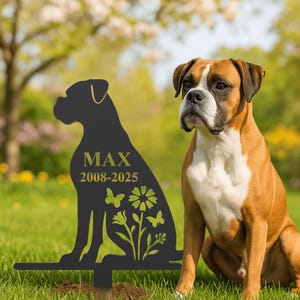Könnte beinhalten: Ein schwarzer Metall-Gartenstecker in Form einer Boxer-Hund-Silhouette, mit dem Namen "MAX" und den Daten "2008-2025" in goldener Schrift. Der Stecker hat Blumen- und Schmetterlingsausschnitte. Ein echter Boxer-Hund sitzt daneben.