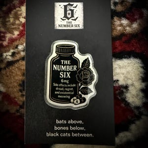 May include: A black card with a white and black graphic of a pill bottle that reads "THE NUMBER SIX" with a rose and pills. The card also has the text "bats above, bones below, black cats between."