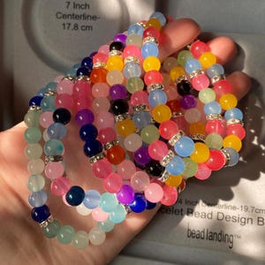 Peut inclure: Une collection de bracelets de perles multicolores composés de perles lisses, rondes et translucides dans des tons de rose, bleu, jaune, vert et noir, agrémentés de petites entretoises argentées avec des cristaux clairs incrustés.