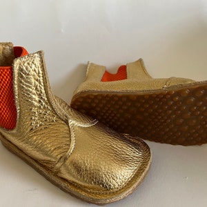 May include: A pair of gold leather ankle boots with orange elastic panels. The boots have a brown rubber sole with a textured pattern.