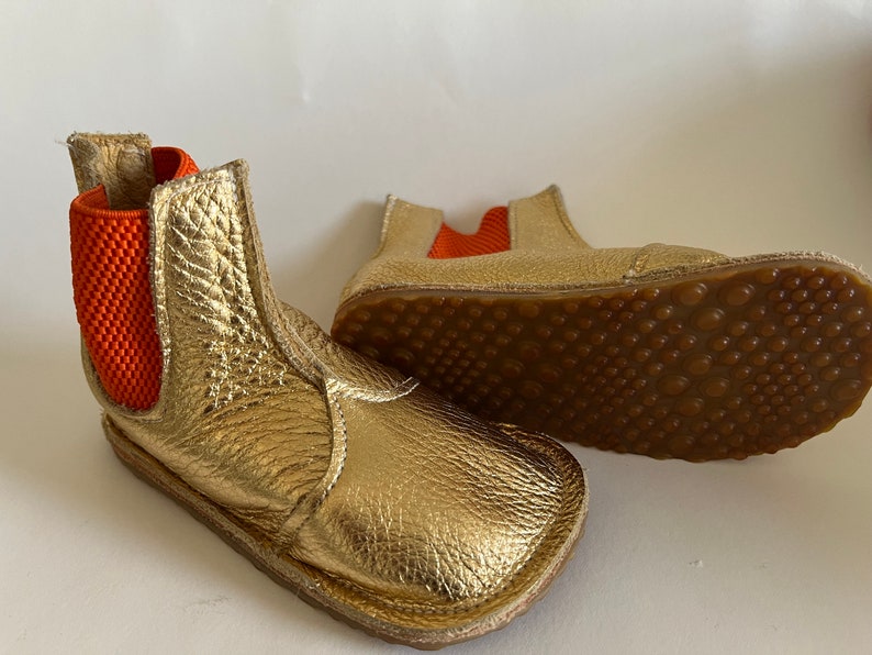 May include: A pair of gold leather ankle boots with orange elastic panels. The boots have a brown rubber sole with a textured pattern.