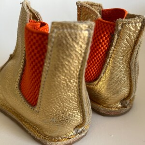 May include: A pair of gold leather ankle boots with orange elastic panels. The boots have a rounded toe and a low heel.