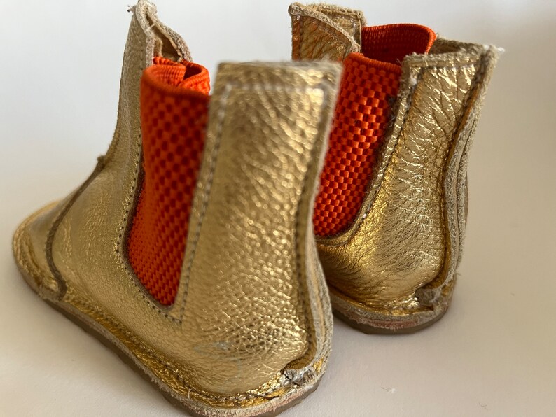 May include: A pair of gold leather ankle boots with orange elastic panels. The boots have a rounded toe and a low heel.