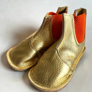 May include: Gold leather ankle boots with orange elastic panels. The boots have a rounded toe and a flat sole.
