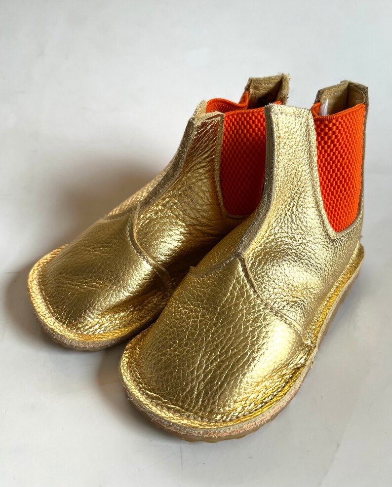 May include: Gold leather ankle boots with orange elastic panels. The boots have a rounded toe and a flat sole.