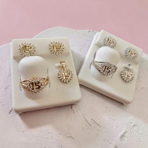 May include: A collection of gold and silver-toned jewelry sets featuring heart-shaped designs with the number 15 at the center. Each piece is accented with small clear stones. The set includes rings, stud earrings, and a pendant on display stands.