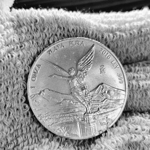 May include: A silver coin with the inscription "1 ONZA PLATA PURA 2016 LEY .999" and a depiction of an angel with wings spread.