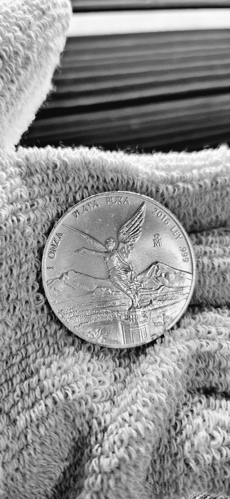 May include: A silver coin with the inscription "1 ONZA PLATA PURA 2016 LEY .999" and a depiction of an angel with wings spread.