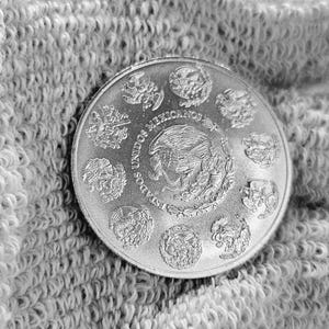 May include: Silver coin with the words "Estados Unidos Mexicanos" and a depiction of the Mexican coat of arms featuring an eagle perched on a cactus with a snake in its beak.