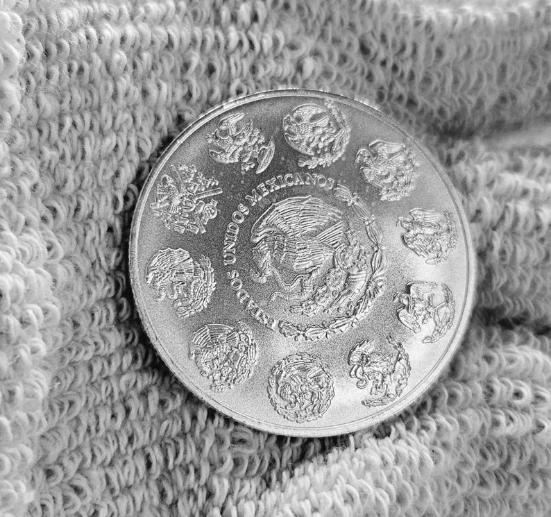 May include: Silver coin with the words "Estados Unidos Mexicanos" and a depiction of the Mexican coat of arms featuring an eagle perched on a cactus with a snake in its beak.
