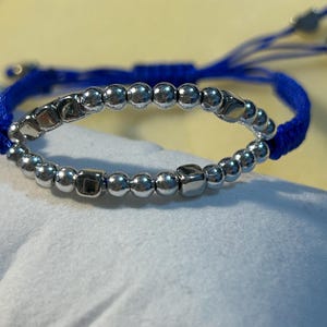 May include: A silver bracelet with a blue cord. The bracelet features a series of small, round silver beads interspersed with square silver beads. The blue cord is adjustable.