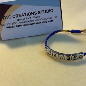 Puede incluir: Una pulsera azul con cuentas de letras plateadas que deletrean "DAWGS" y símbolos de corazón. La pulsera está sobre una superficie amarilla, con una tarjeta de visita de DTC Creations Studio en el fondo.