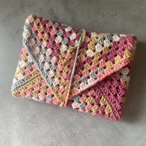 May include: A crocheted blanket with a geometric pattern in shades of pink, gray, and beige. The blanket is folded in a triangular shape.