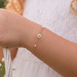 May include: A delicate gold chain bracelet featuring a small flower motif composed of tiny white pearls, with additional single pearls spaced along the chain, worn on a wrist while holding a white woven basket filled with small white flowers.