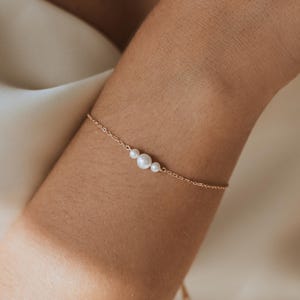 May include: A delicate gold bracelet featuring three white pearls. The bracelet is worn on a wrist, with a soft, neutral-toned background. The pearls are of varying sizes, adding a subtle elegance to the jewelry.