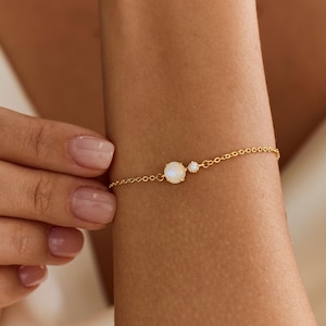 Op de afbeelding: Een delicate gouden kettingarmband met een ronde, iriserende witte edelsteen en een kleinere, heldere ronde steen gezet in goud. De armband is voorzien van een fijne schakelketting en heeft een minimalistisch ontwerp dat op de huid rust.