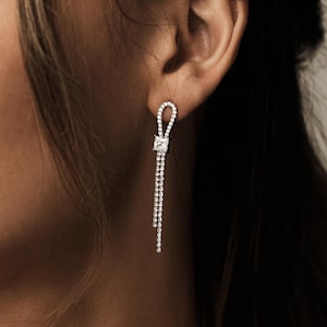 Op de afbeelding: Elegante zilveren oorbellen met een vierkant geslepen middensteen en een bungelende ketting van kleinere stenen. De oorbellen zijn ontworpen met een lusvormige bovenkant en worden getoond op een oor, wat hun delicate en verfijnde ontwerp benadrukt.