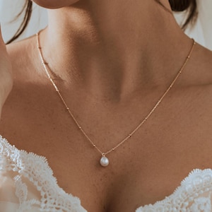 Peut inclure: Un collier délicat en or avec un pendentif en perle unique. Le collier est porté par une personne avec un haut en dentelle blanche et un voile.