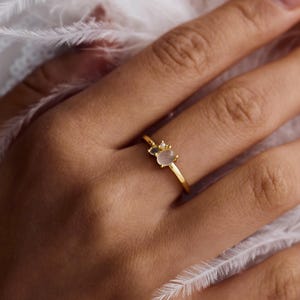 May include: A gold ring with a small, light pink gemstone and a smaller clear stone. The ring is on a person's finger, with white feathers in the background. The ring is a delicate piece of jewelry.